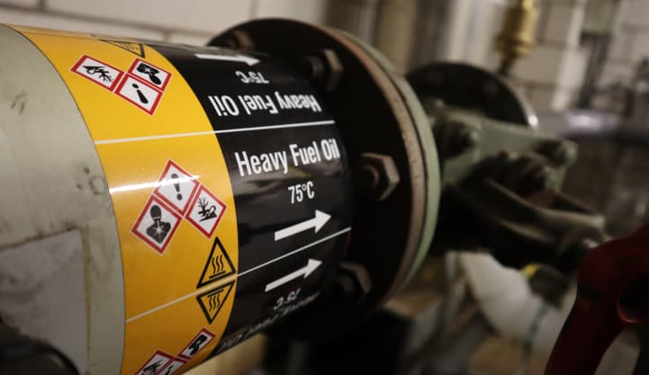 Industrial pipeline labeled with a Heavy Fuel Oil pipe marker featuring GHS hazard symbols and directional flow indicators